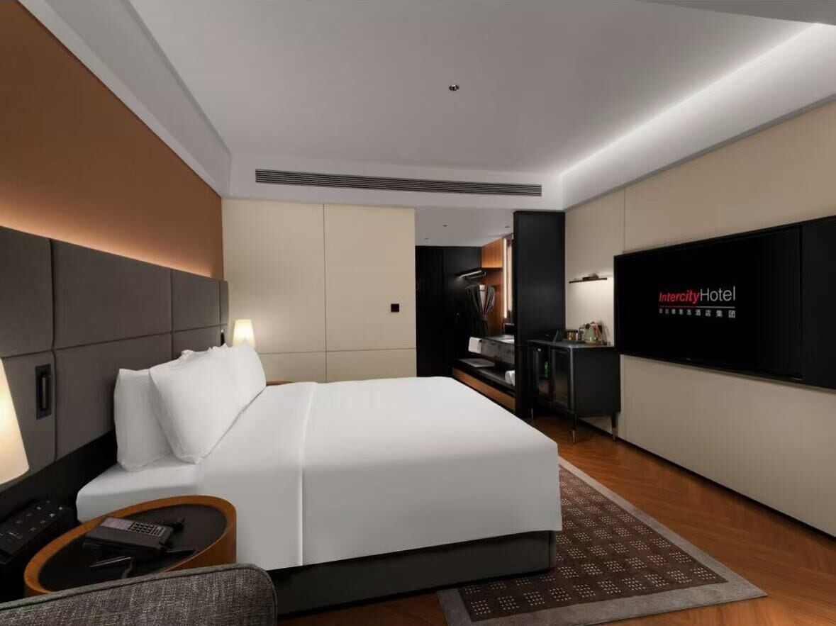A modern hotel room with a large bed, a desk, a television, and a kitchenette.