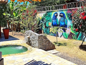 Ein Steinkunstbrunnen, eine Wandmalerei mit Papageien und Kübelpflanzen in einem Garten.