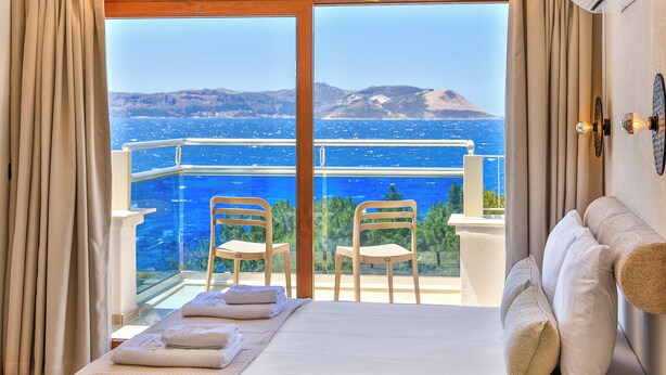 A hotel room with a bed, two chairs, a balcony with a view of the sea and mountains, and a window with curtains.