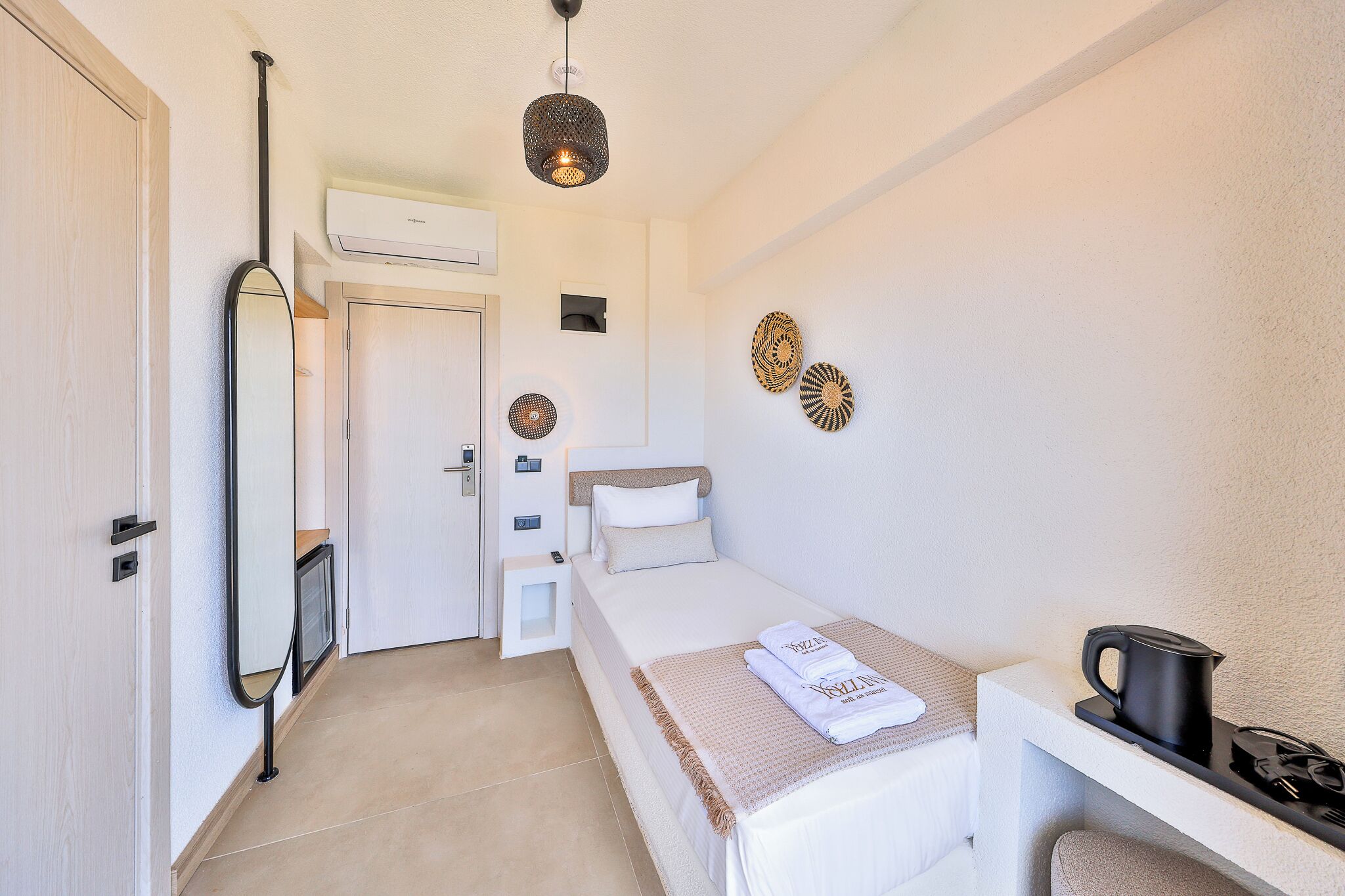 A small, clean room with a bed, a desk, a kettle, and a mirror.
