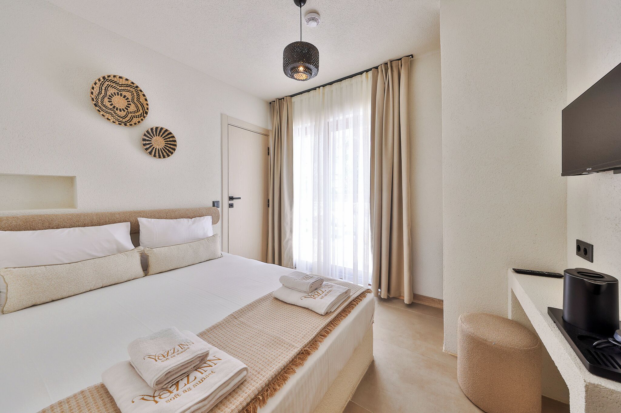 A hotel room with a bed, a desk with a coffee machine, a TV, and a large window with curtains.