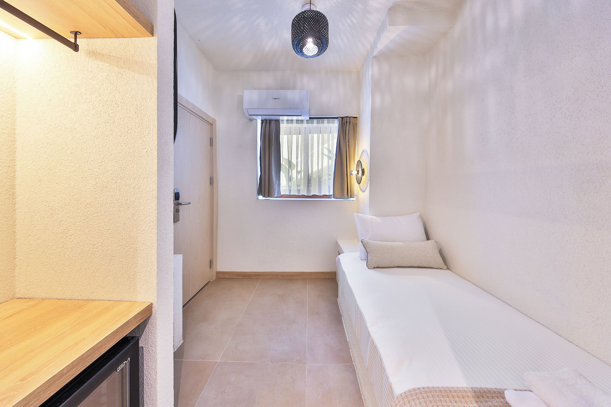 Een kleine kamer met een bed, een raam met gordijnen, een airconditioning en een plafondlamp.