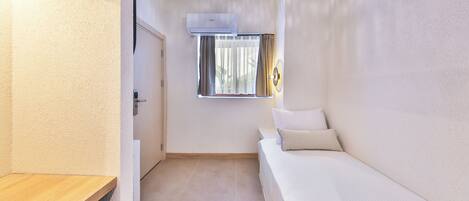 A compact room with a bed, a window with curtains, an air conditioning unit, and a ceiling light fixture.