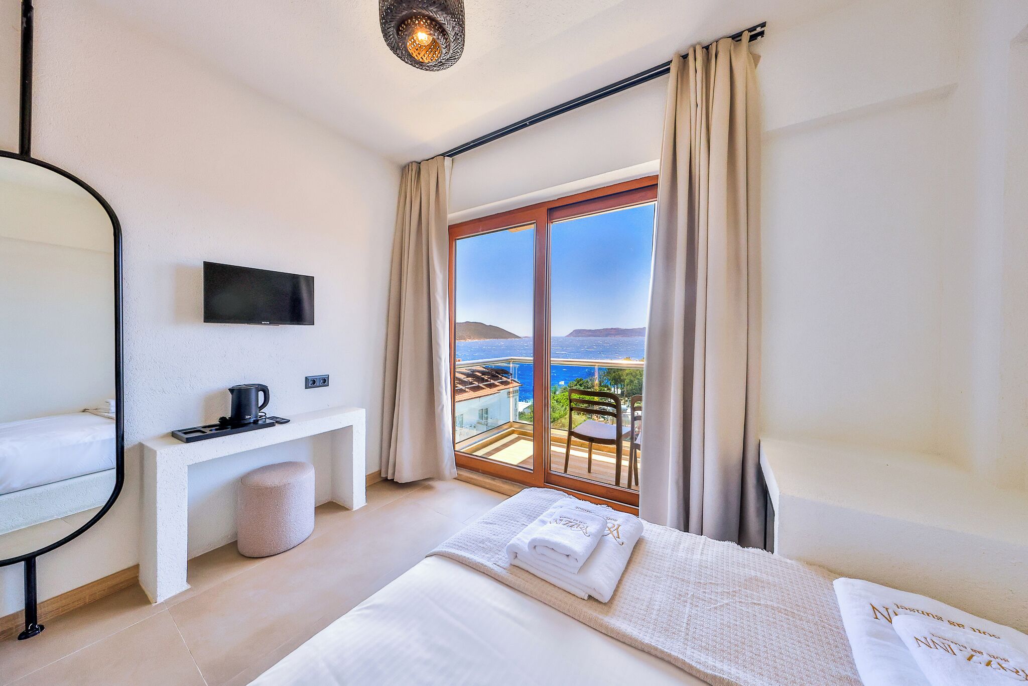 A hotel room with a bed, a TV, a desk with a coffee maker, a small stool, and a view of the sea.