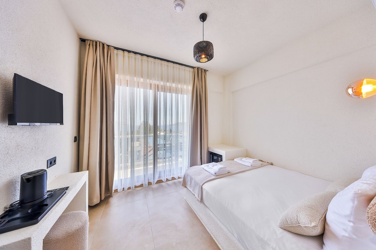 A hotel room with a bed, a TV, a desk with a kettle, and a window with curtains.