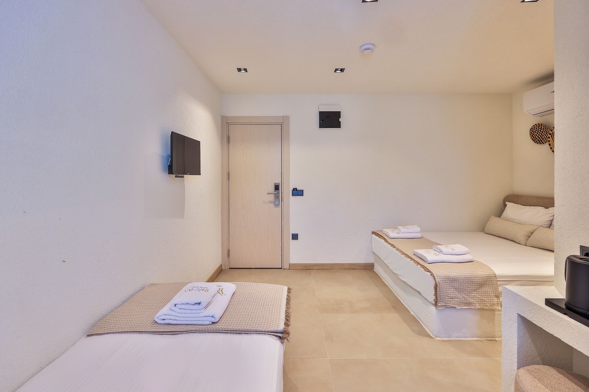 A hotel room with two beds, a TV, a door, and a small desk with a kettle.