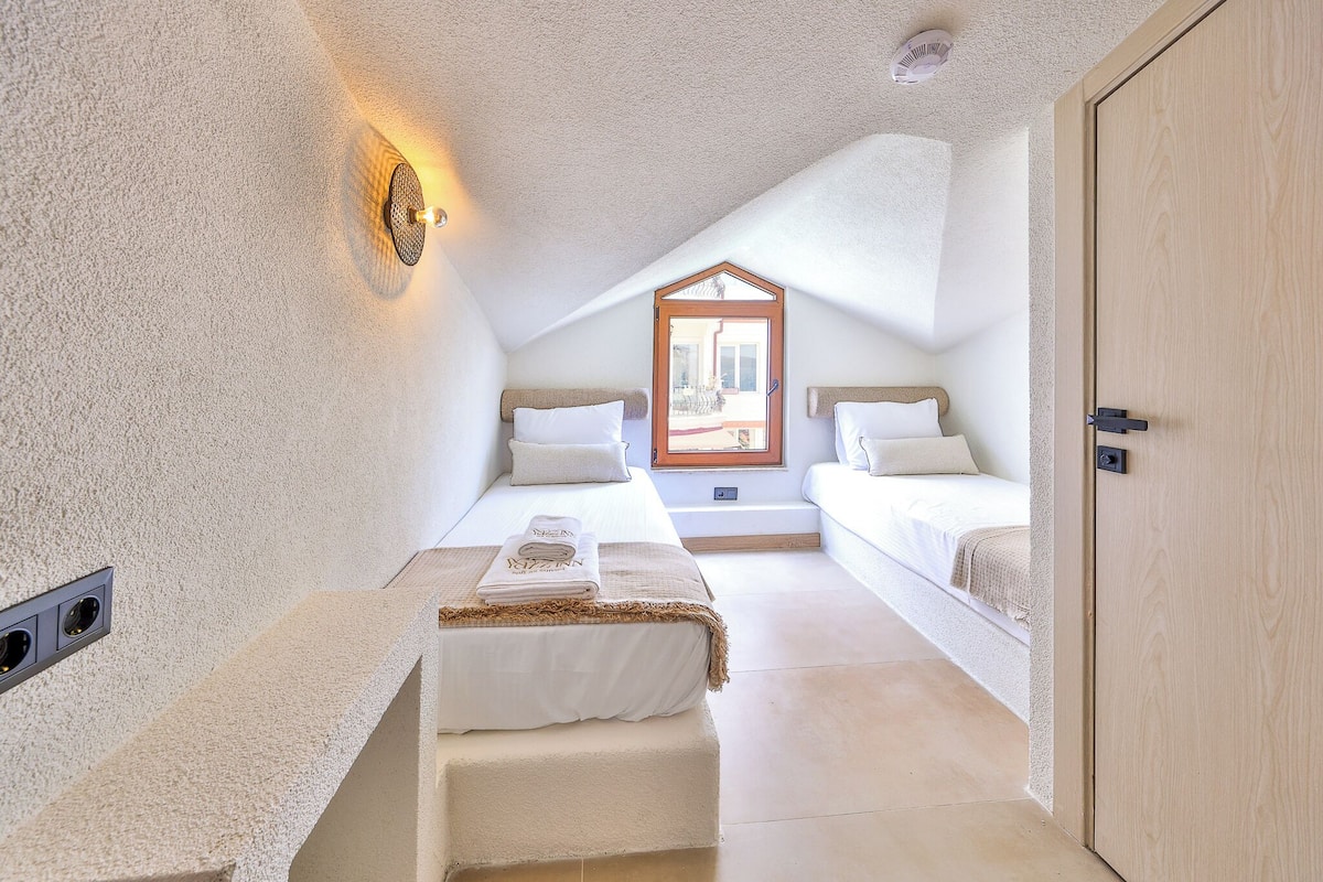 A small, cozy room with a sloped ceiling, a bed with a headboard, a bedside table, and a window with a view.