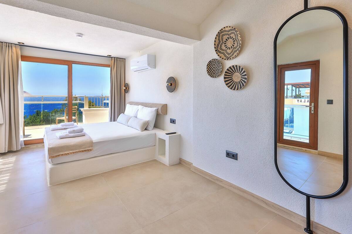 A bedroom with a bed, a nightstand, a mirror, and a view of the sea.