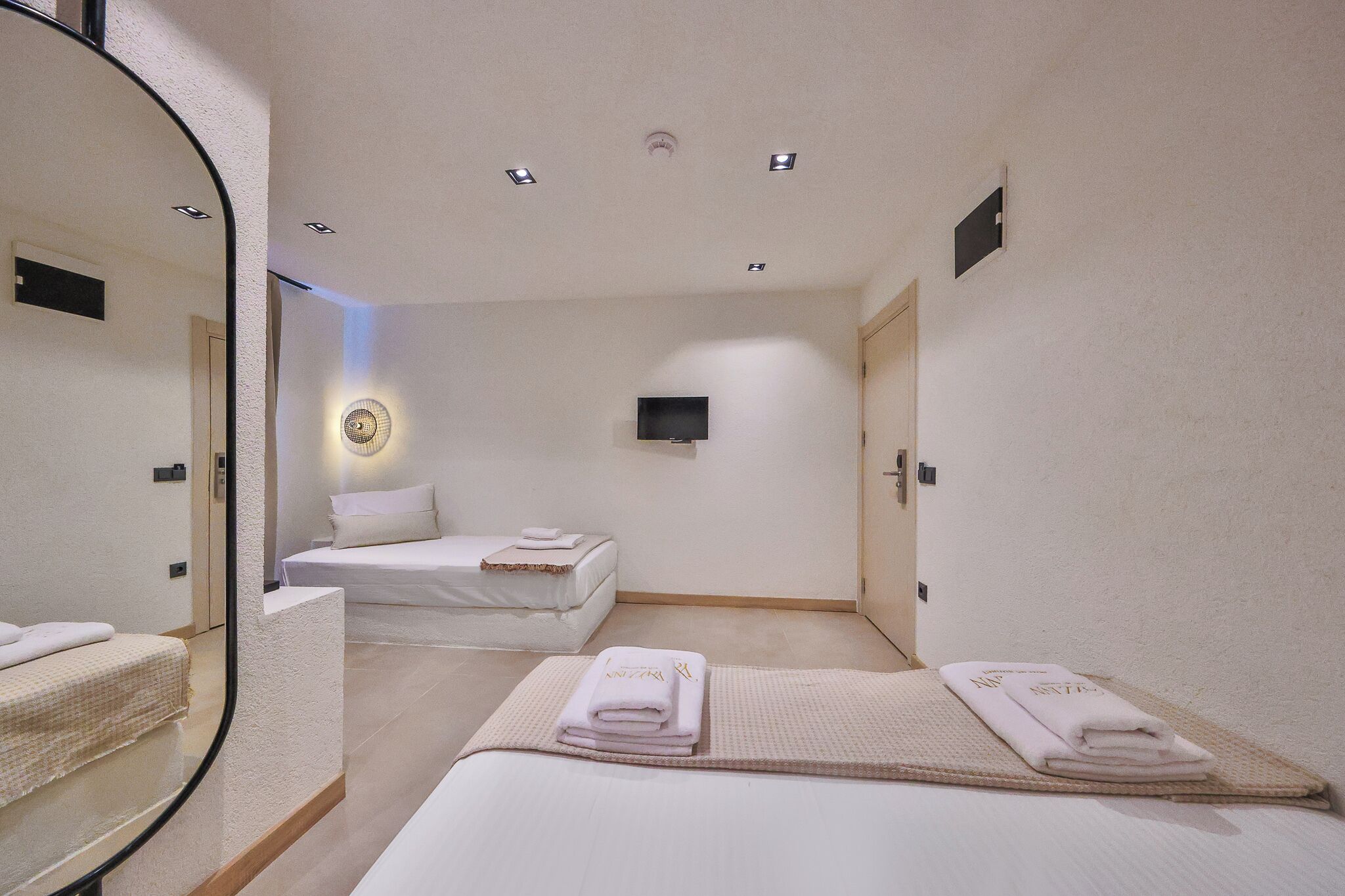 A hotel room with two beds, a mirror, a wall-mounted TV, and a ceiling light.