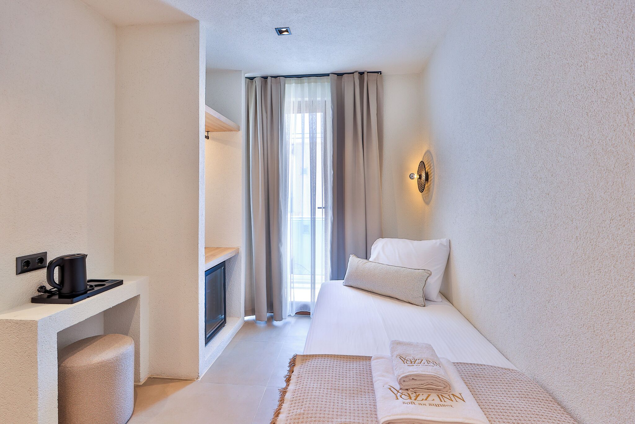Une chambre d’hôtel avec un lit, un bureau doté d’une bouilloire, une lampe fixée au mur et une fenêtre avec des rideaux.