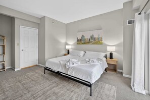 Un dormitorio con cama, dos mesitas de noche, un cuadro de paisaje urbano, una puerta blanca y una ventana con cortina.