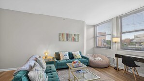 Ein modernes Wohnzimmer mit einem türkisfarbenen Sofa, einem Glastisch, einem Sitzsack und einem Schreibtisch mit Stuhl.