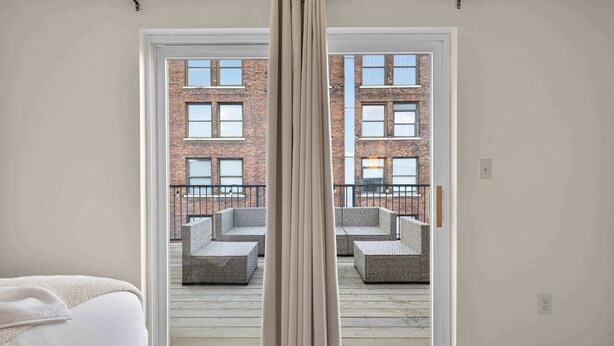 Ein Hotelzimmer mit Bett, Vorhang und Blick auf einen Außenbalkon mit Sitzgelegenheit.