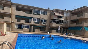 Ein Schwimmbecken mit gefliestem Boden, umgeben von einem Gebäude mit mehreren Balkonen und blauem Himmel.