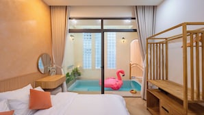 A bedroom with a bed, a wooden bench, a small table, a mirror, a pink flamingo float, and a view of a swimming pool. - Hometown Nimi 3 BRS Pool Villa Da Nang (Da Nang)