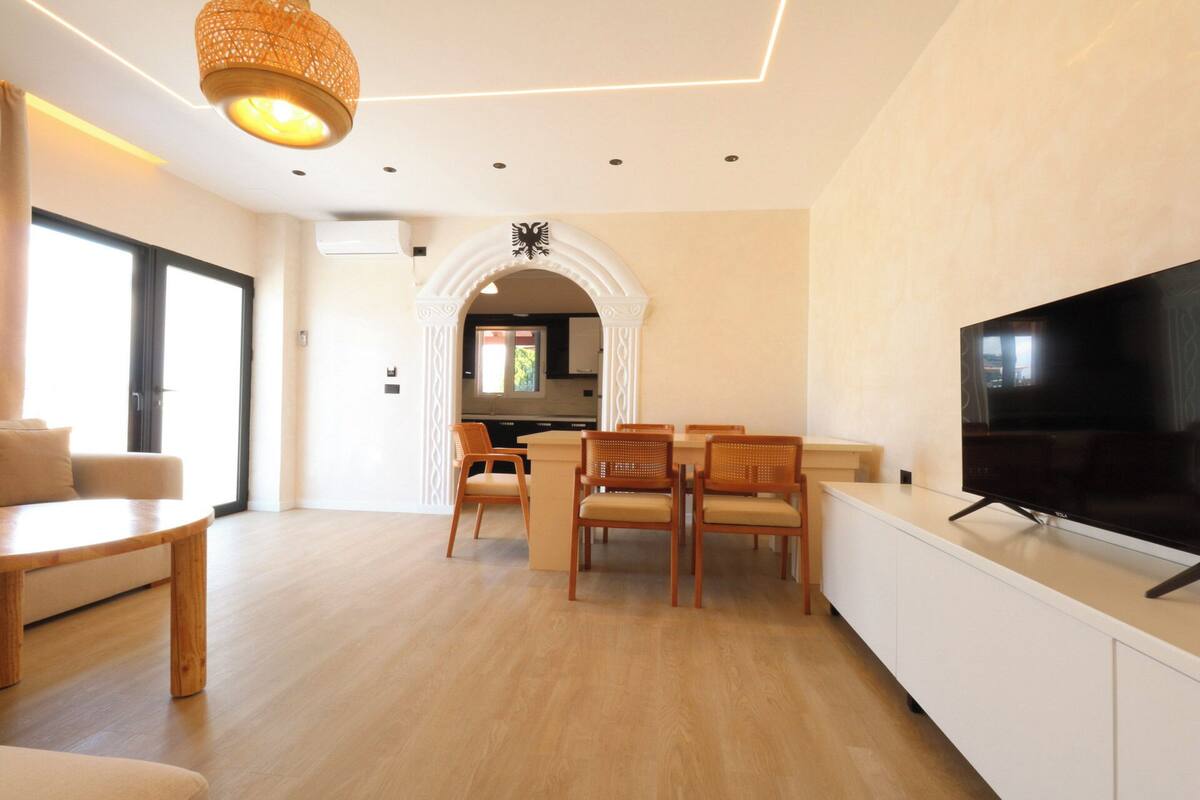 A modern living room with a TV, wooden furniture, and a kitchen in the background.