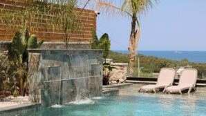 A pool with a waterfall feature, lounge chairs, and a view of the ocean. - Luxury Villa Oasis (Ksamil)