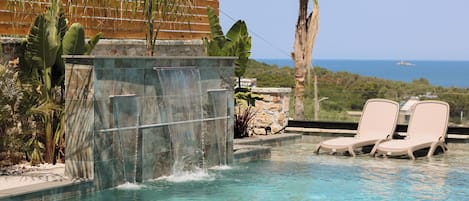 A pool with a waterfall feature, lounge chairs, and a view of the ocean.