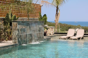 A pool with a waterfall feature, lounge chairs, and a view of the ocean. - Luxury Villa Oasis (Ksamil)