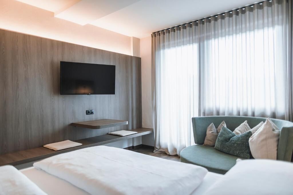 Habitación de hotel moderna con un televisor de pantalla plana montado en una pared de paneles de madera, una mesita de noche con lámpara, un sofá verde con cojines y un ventanal con cortinas de gasa.