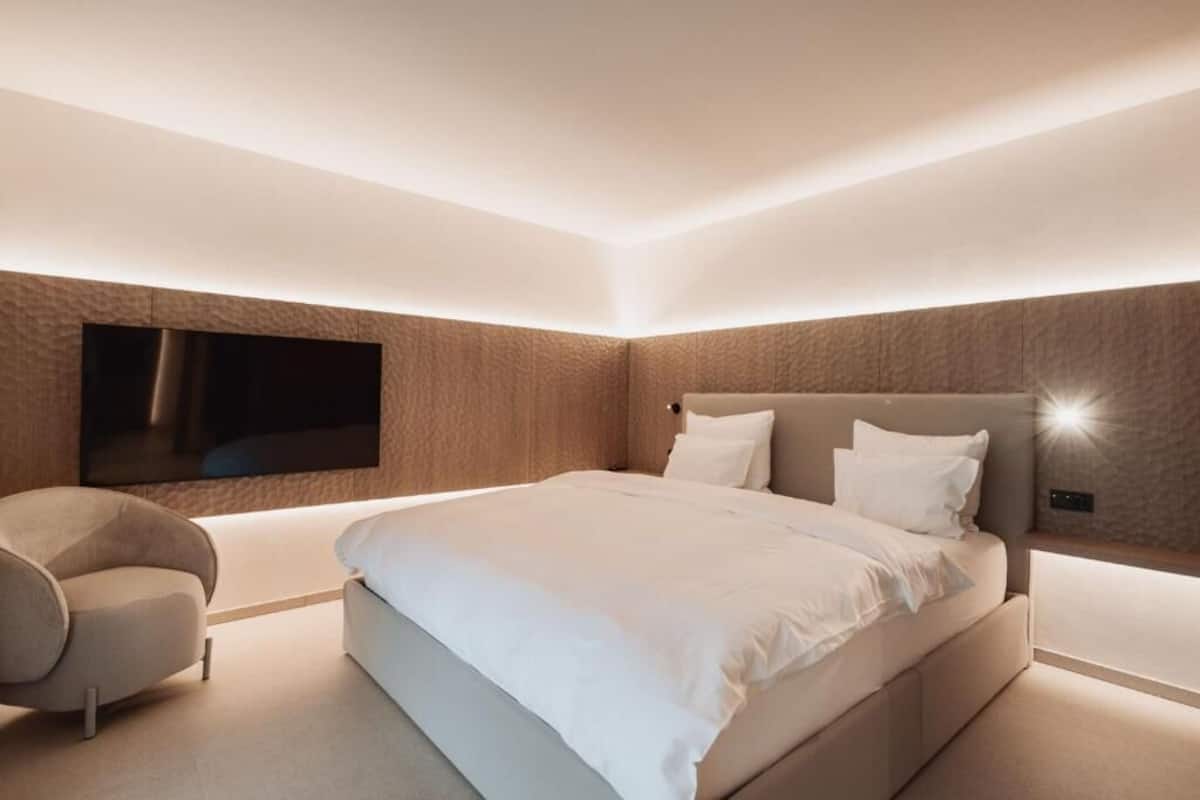 A modern bedroom with a large bed, a flat-screen TV mounted on the wall, and a unique curved chair.
