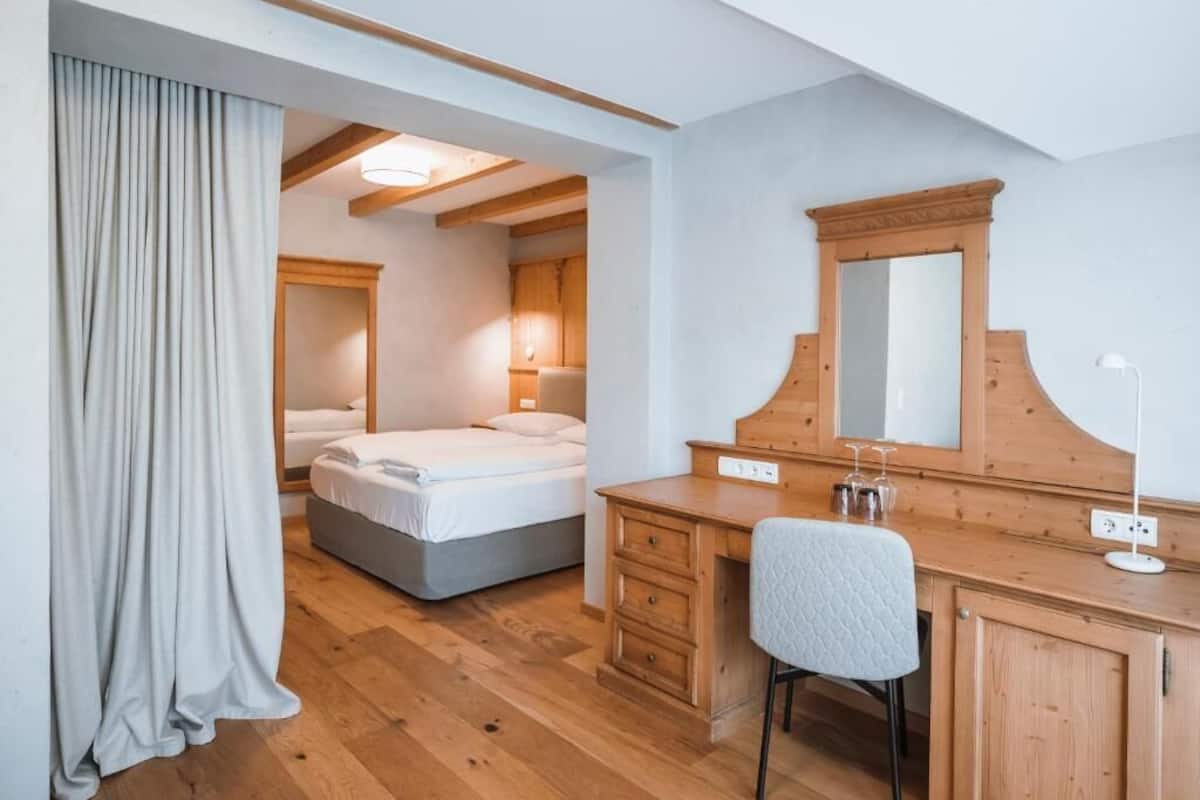 A bedroom with a bed, a wooden desk, a chair, a mirror, and a window.