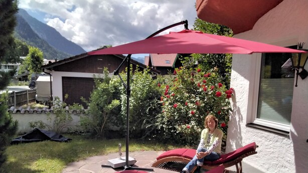 Eine Terrasse mit einem roten Sonnenschirm, einer Liege und einem Garten mit roten Blumen.