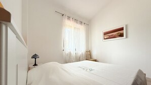 Ein Schlafzimmer mit Bett, Nachttisch, Lampe, Stuhl, Fenster mit Vorhängen und einem gerahmten Bild an der Wand.