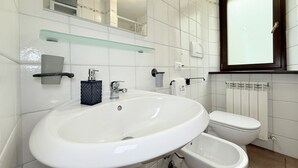 Ein modernes Badezimmer mit weißem Waschbecken, WC und Bidet, einer Glasablage über dem Waschbecken sowie einem Spiegel mit dunkler Umrandung.