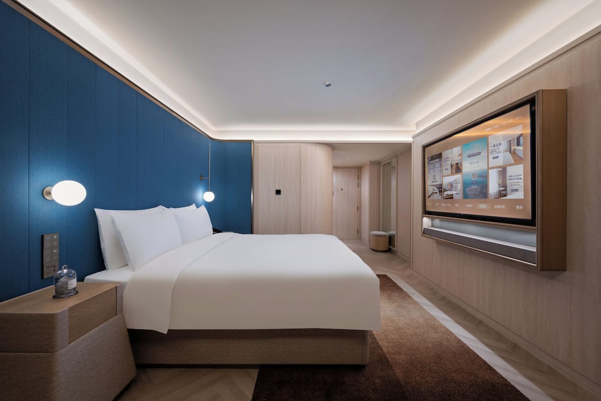 A modern hotel room with a large bed, a flat-screen TV mounted on the wall, and a nightstand with a lamp.