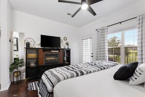 A bedroom with a fireplace, a bed with patterned bedding, a television on a stand, and a view of the outdoors through a window. - FW15, Prime Golf View 1BR - No Stairs! Walk to Pool & Amenities (Branson West)