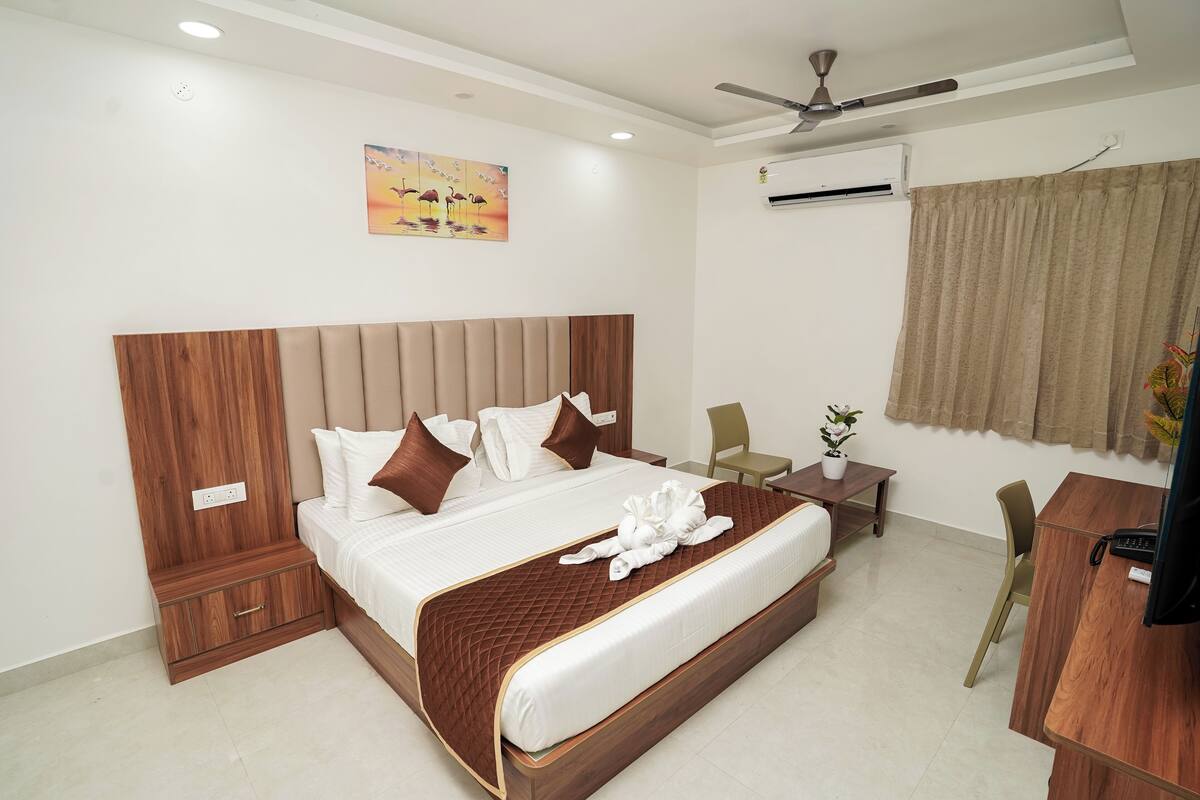 A hotel room with a large bed, a desk, a chair, a small table with a plant, and an air conditioning unit.