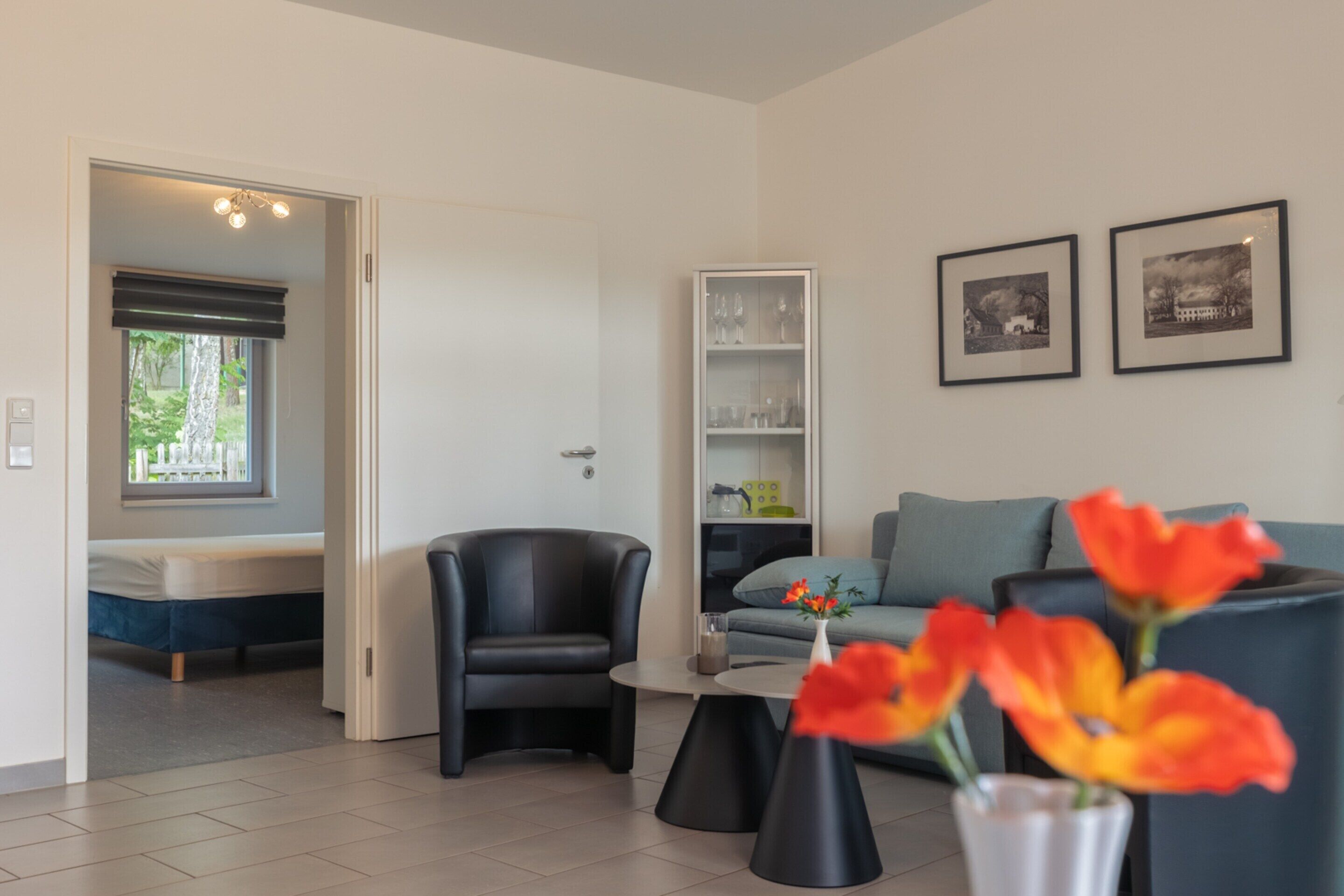 A modern living room with a sofa, a chair, a coffee table, and a vase with flowers. There is a bed in the background and a door leading to another room.