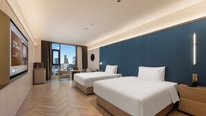 Ein Hotelzimmer mit zwei Betten, einem Schreibtisch, einem Stuhl, einem Fernseher und Blick auf die Stadt.