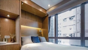 Un dormitorio moderno con una cama grande, cabecera de madera, mesitas de noche y vistas de la ciudad a través de amplios ventanales.