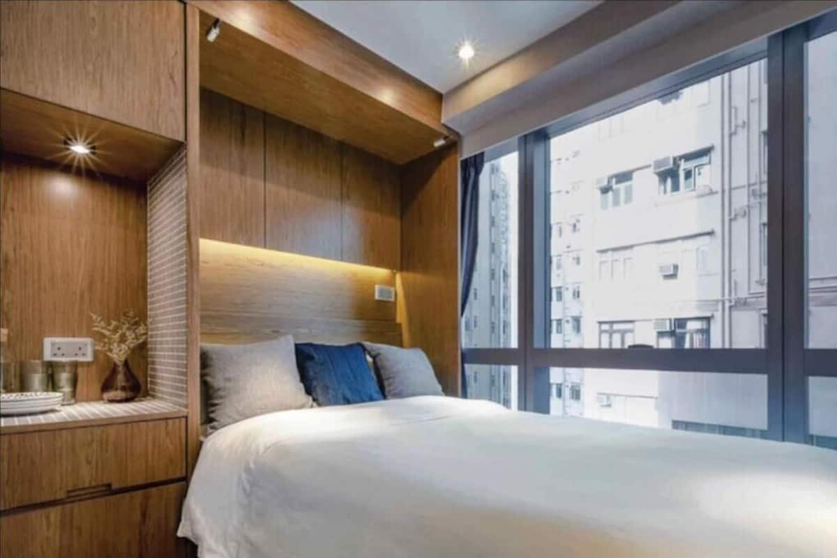A modern bedroom with a large bed, wooden headboard, bedside tables, and a view of the city through large windows.