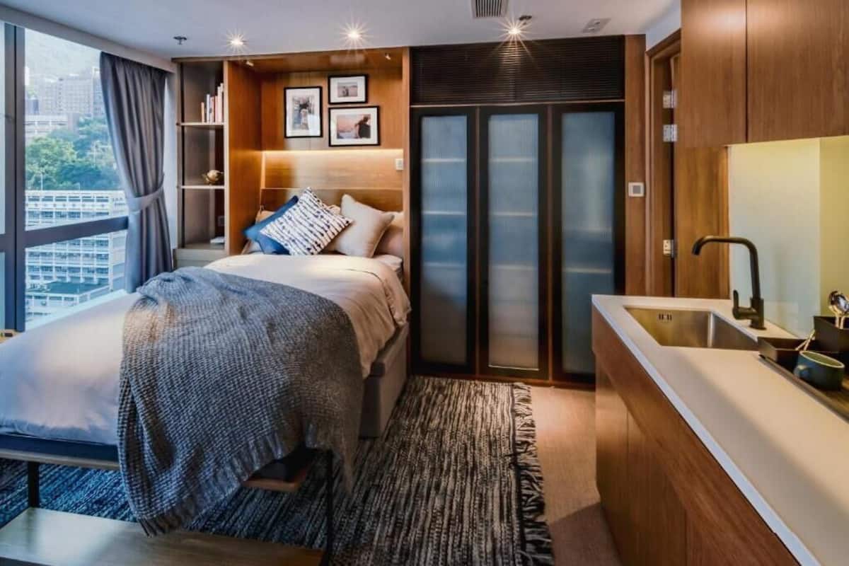 A modern hotel room with a bed, bedside tables, a built-in wardrobe, a large window, and a bathroom with a sink and mirror.