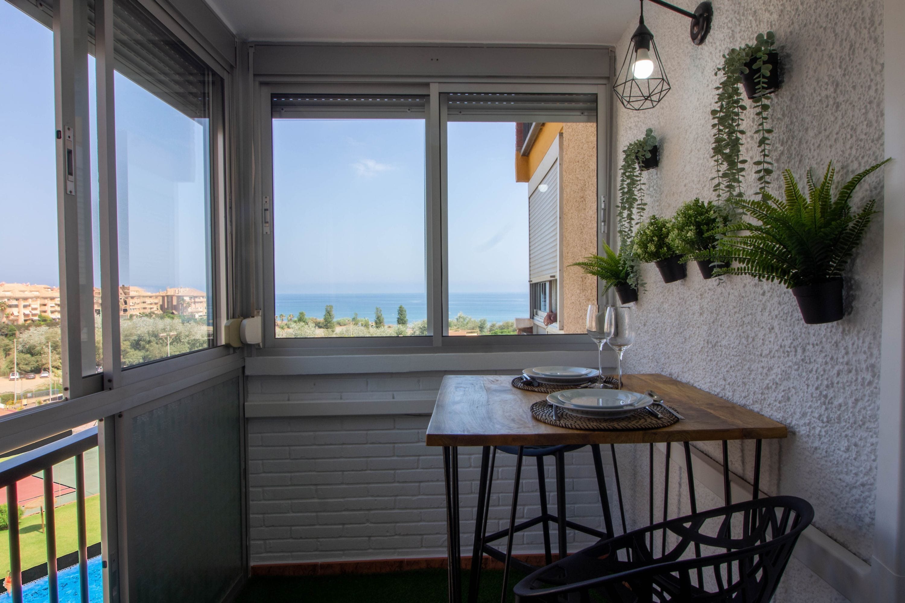 A balcony with a view of the sea, a table with plates and a glass, and potted plants on the wall.