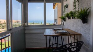 Un balcón con vista al mar, una mesa con platos y una copa, y plantas en macetas en la pared.