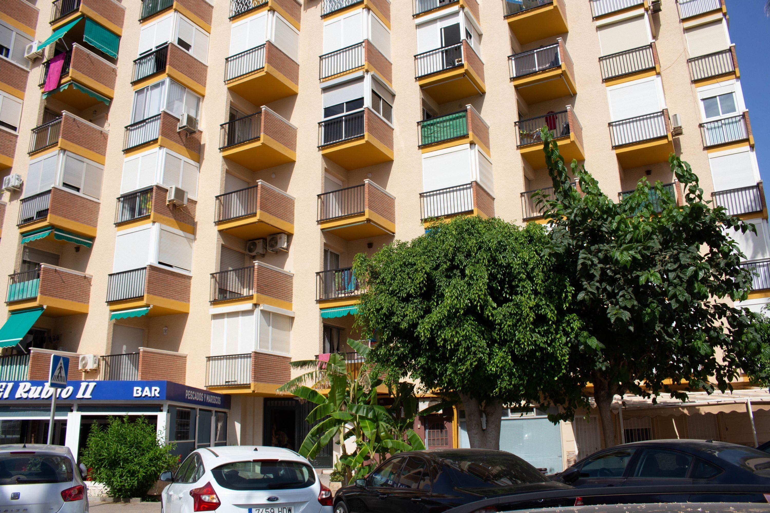 Un immeuble à plusieurs étages doté de balcons, un bar appelé « El Rubio II » et des voitures garées.