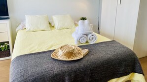 Een net opgemaakt bed met kussens, een bedlampje, een opgerolde handdoek, een hoed en een kleine plant op het bed.