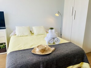 Una cama bien hecha con almohadas, una lámpara de noche, una toalla enrollada, un sombrero y una planta pequeña sobre la cama.