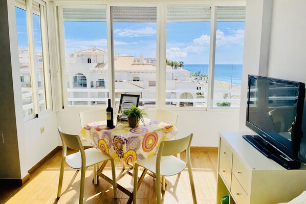 A dining area with a table set for two, a television on a cabinet, and a view of the sea through large windows. - Capri 403 by At Home Costa del Sol (Torrox)