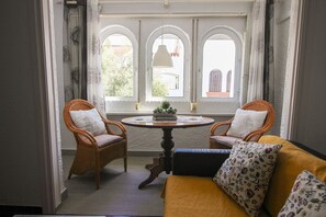 Una habitación con una mesa redonda, dos sillones de mimbre, un sofá y tres ventanas arqueadas que dan al exterior.