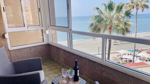 En balkon med fletstole, et lille bord med en flaske og glas, samt udsigt over stranden og havet.