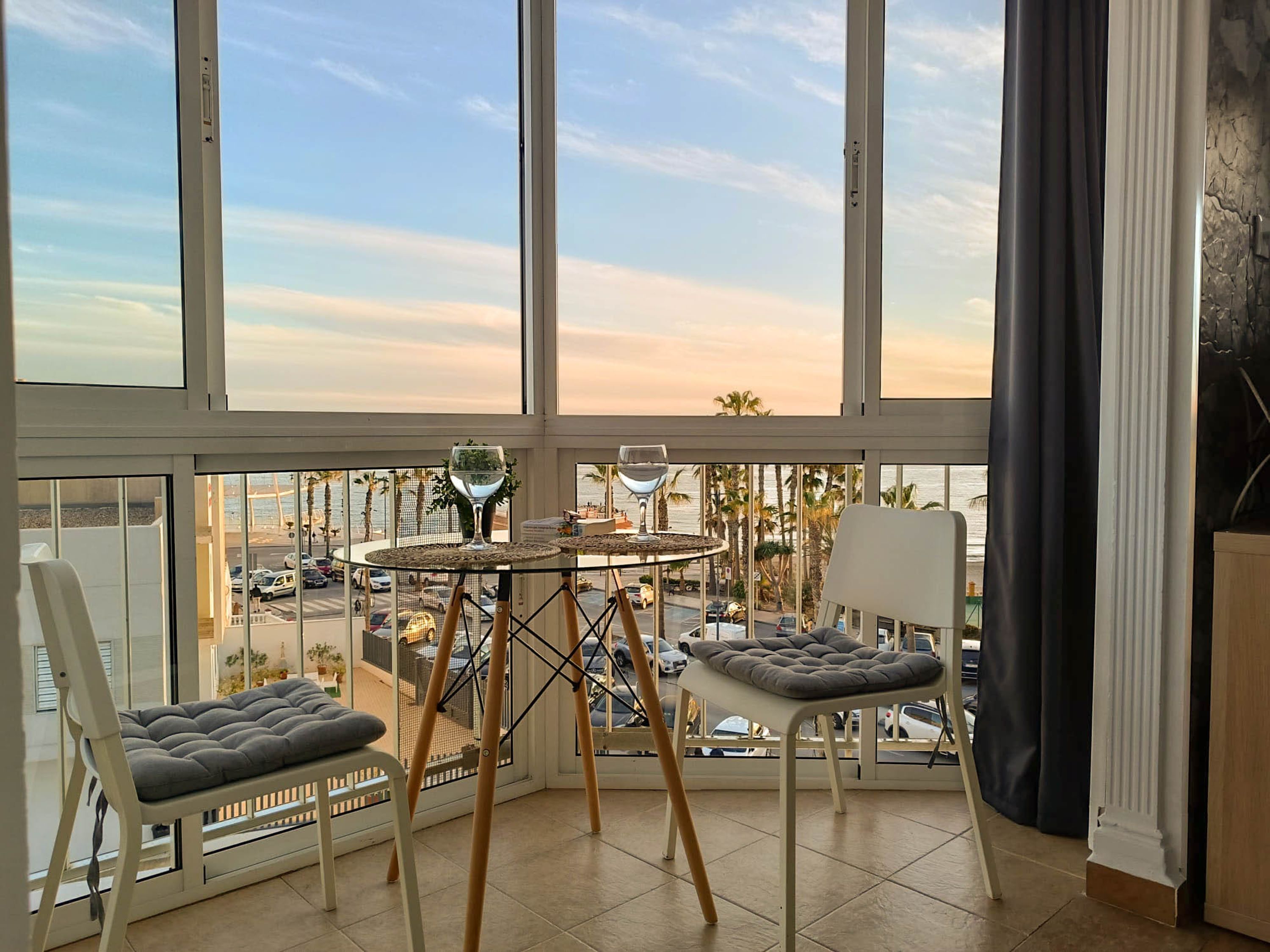 A balcony with a glass table, two cushioned chairs, and a view of the street and palm trees.