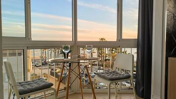 A balcony with a glass table, two cushioned chairs, and a view of the street and palm trees.