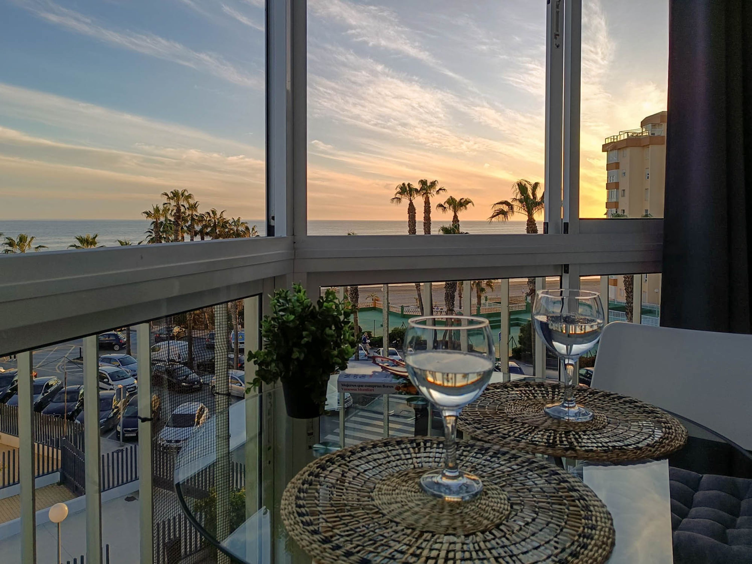 A balcony with a view of the ocean and palm trees, a glass table with a woven centerpiece, and a potted plant.