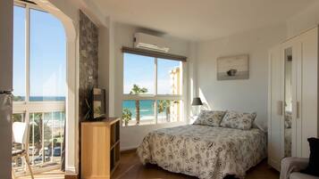 A bedroom with a bed, a TV on a stand, a window with a view of the sea, and a balcony.