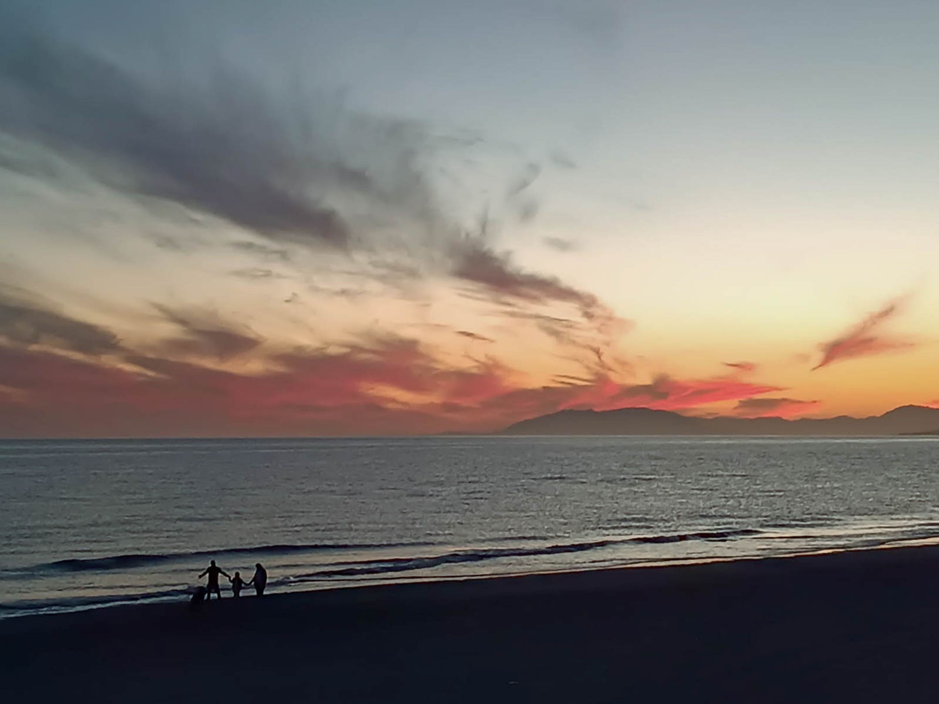 A beach scene with a sunset, calm sea, and silhouettes of two people walking.